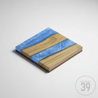 Square wooden coaster with a blue resin river design. The coaster features two wooden panels with a blue resin river flowing between them. The coaster is approximately 4 inches square and has a cork base.