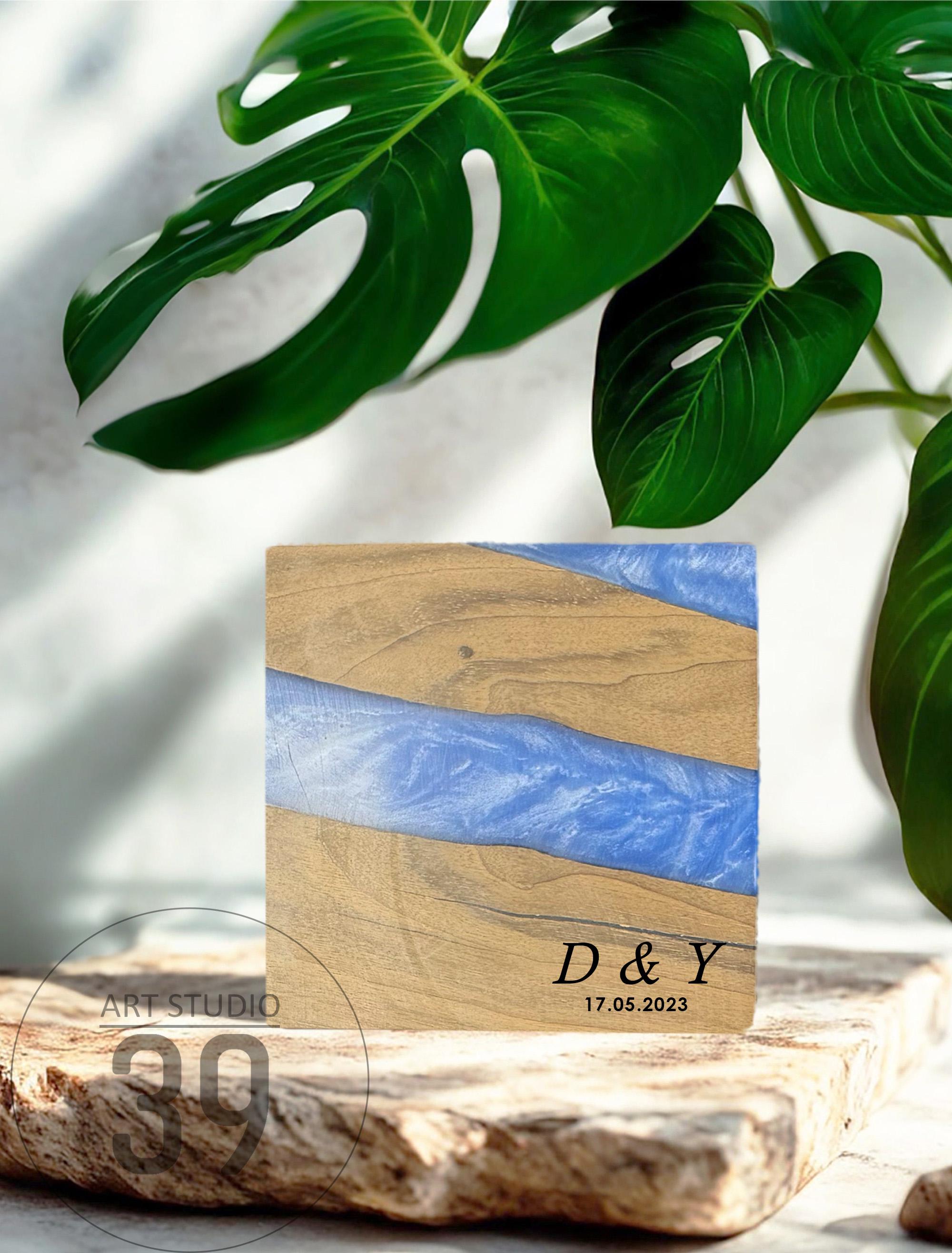 Square wooden coaster with a blue resin river design. The coaster has a natural wood grain pattern and is inscribed with the initials "D &amp; Y" and the date "17.05.2023". The coaster is displayed on a stone surface.