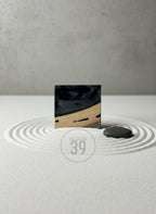 A square, black and wood-toned decorative object with a smooth, glossy finish. The object is set on a bed of white sand with a small, dark gray stone. The image includes the text "ART STUDIO 39".