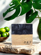 A square, two-toned decorative piece with a black top and a light brown bottom. The bottom section has the engraved initials "D &amp; Y" and the date "17.05.2023". The piece is displayed on a stone surface, with green limes and tropical leaves in the background.