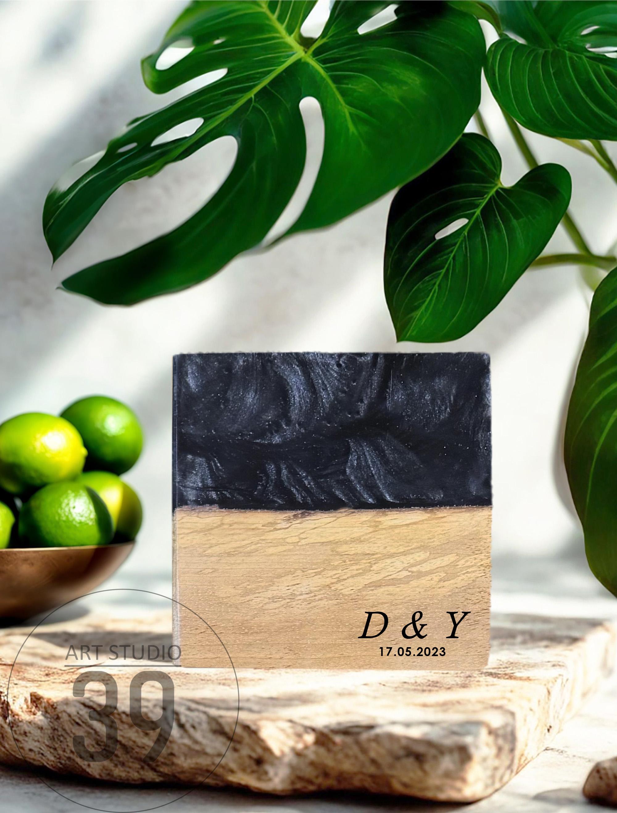 A square, two-toned decorative piece with a black top and a light brown bottom. The bottom section has the engraved initials "D &amp; Y" and the date "17.05.2023". The piece is displayed on a stone surface, with green limes and tropical leaves in the background.