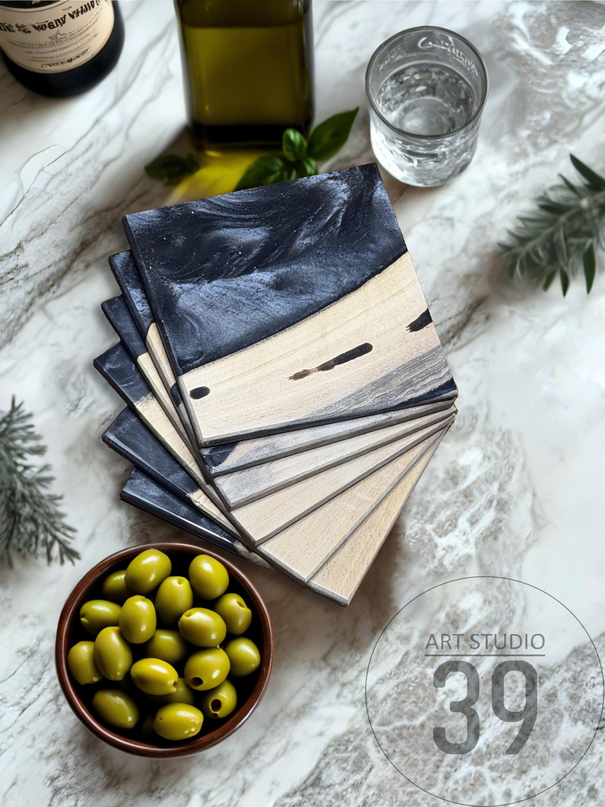 A stack of square coasters with a two-tone design. The top half is a dark, marbled gray, and the bottom half is a light wood grain. A bowl of green olives, a glass of water, and a bottle of olive oil are also visible.