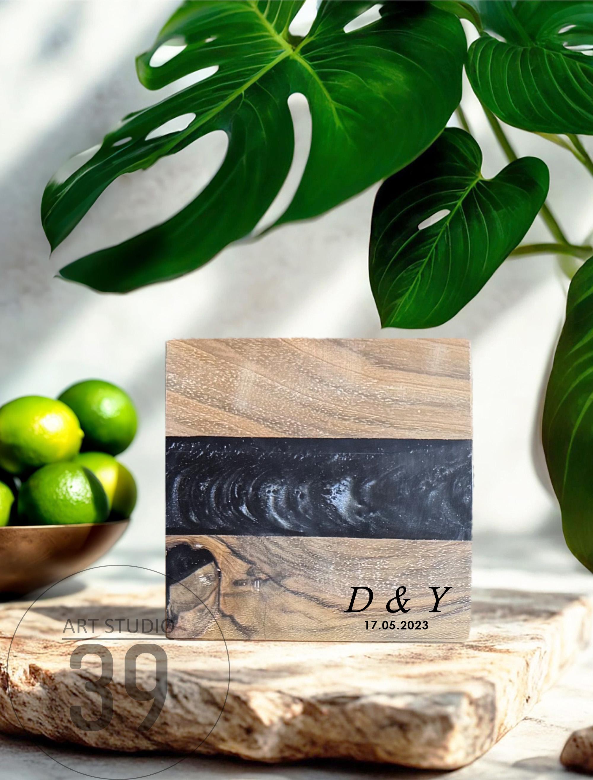 A square wooden block with a black resin stripe. The wood has a natural grain pattern. The block is engraved with the initials "D &amp; Y" and the date "17.05.2023". Limes in a bowl and green leaves are in the background.