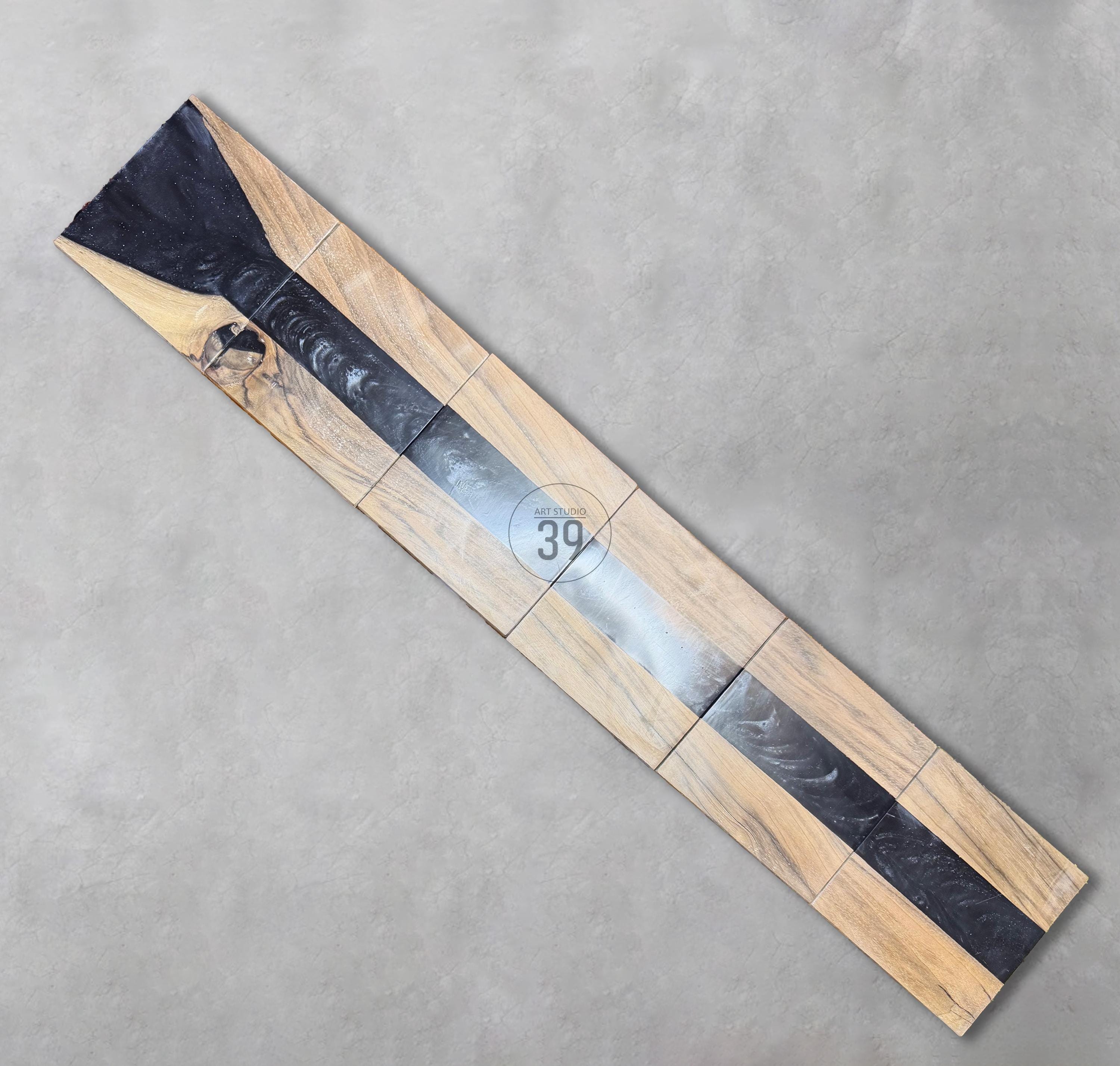 A rectangular wooden table with a dark gray epoxy resin river. The table is made of several wooden blocks with a natural wood grain pattern. The epoxy river runs down the center. The text "ART STUDIO 39" is in the center.