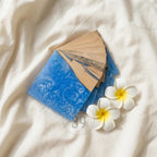 A set of square coasters with a blue resin design and natural wood accents. The coasters are arranged on a white surface, with two white and yellow flowers nearby. The coasters are approximately 4 inches square.