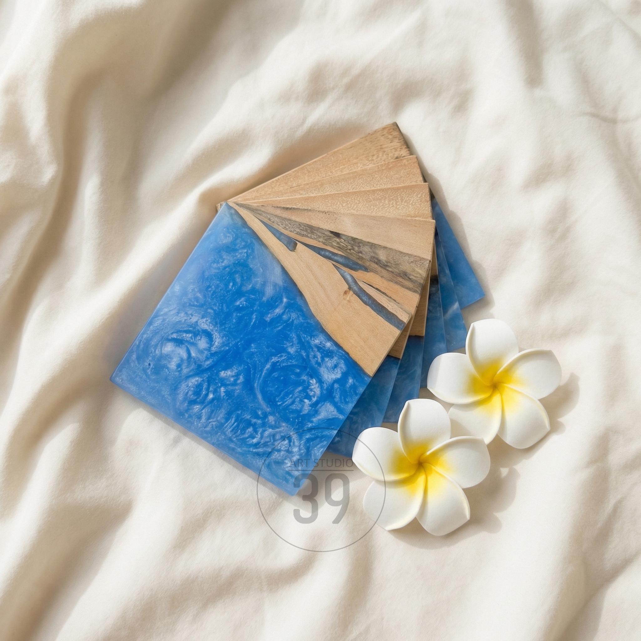 A set of square coasters with a blue resin design and natural wood accents. The coasters are arranged on a white surface, with two white and yellow flowers nearby. The coasters are approximately 4 inches square.