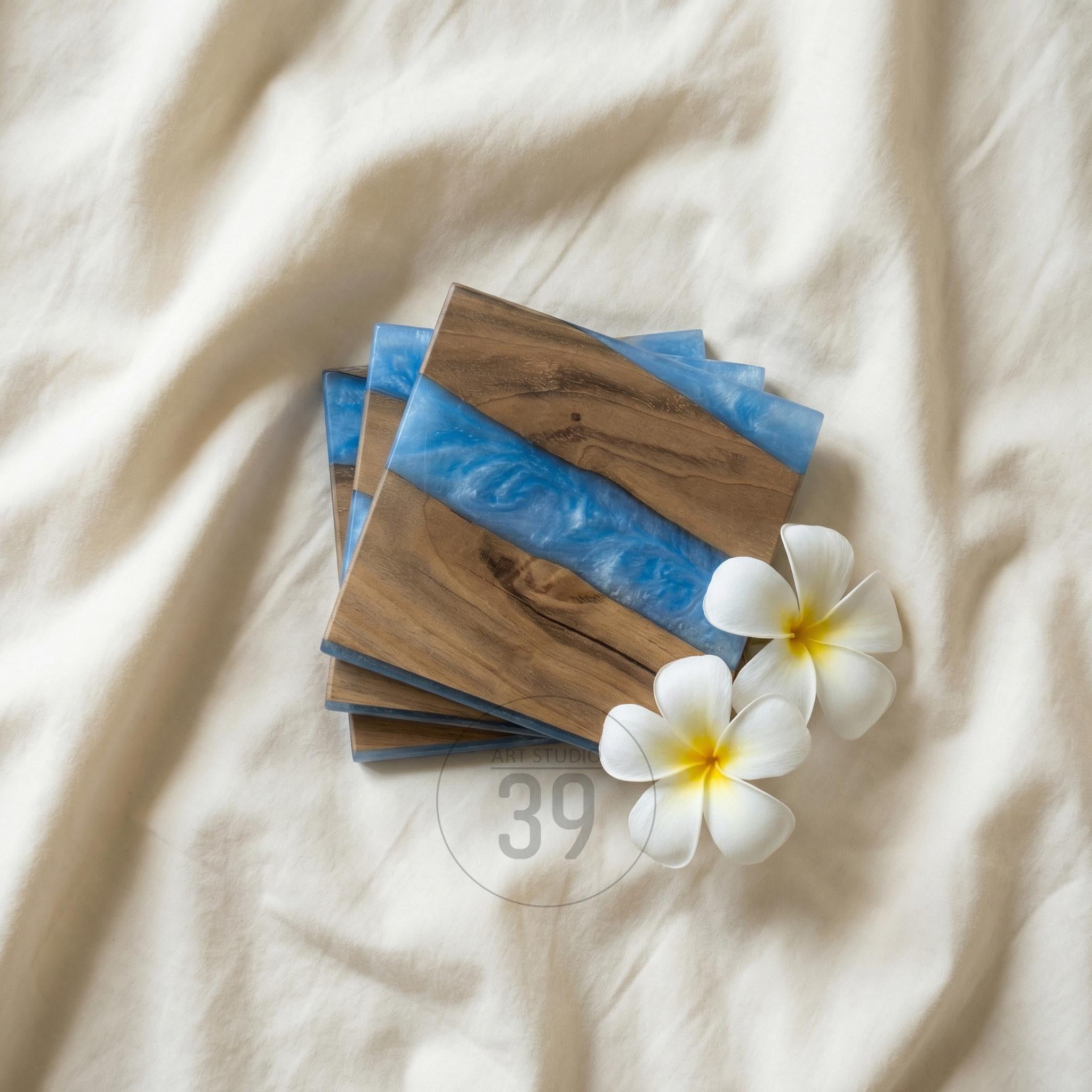 A stack of square wooden coasters with a blue resin stripe. The coasters have a natural wood grain pattern and are accented with white flowers. The number "39" is visible on the bottom right.