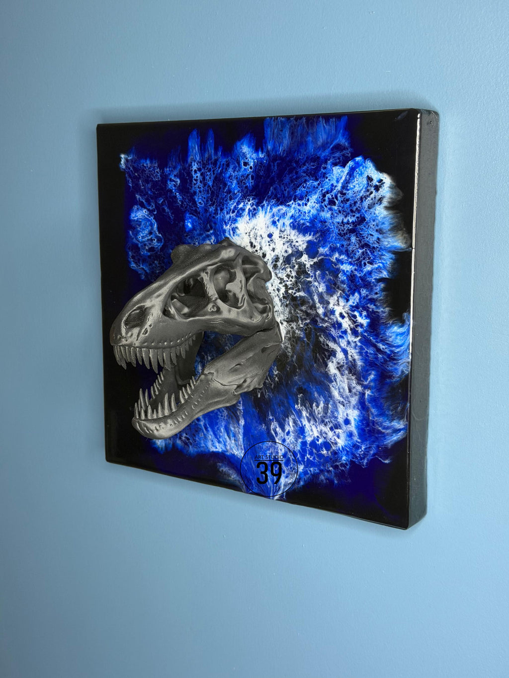 A square wall art piece featuring a metallic gray dinosaur skull against a vibrant blue and white abstract background. The artwork is mounted on a black frame, with the number "39" visible.