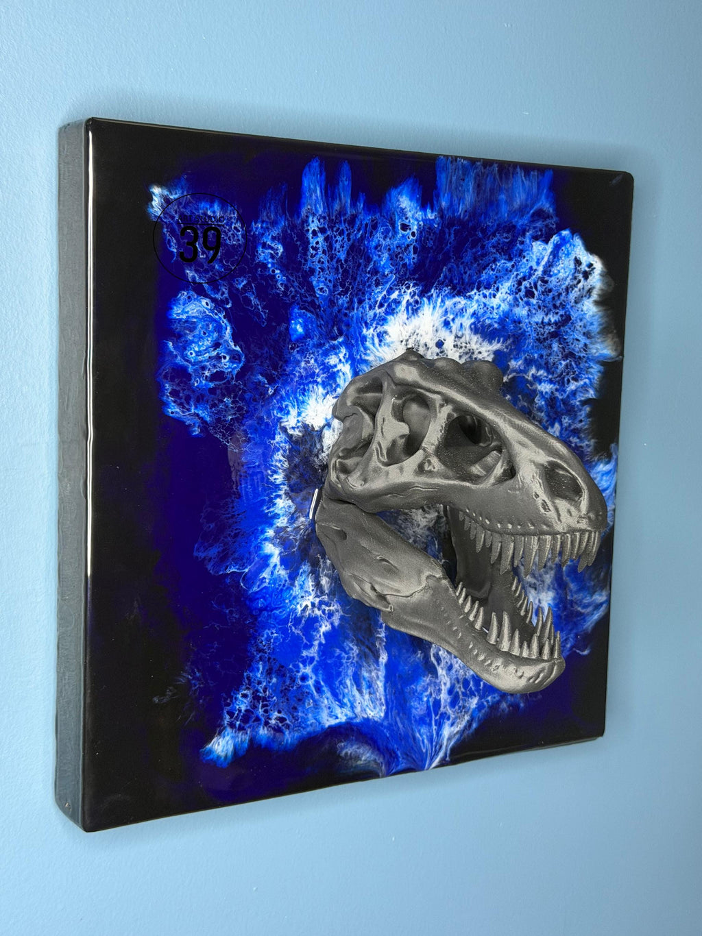 A square wall art piece featuring a gray dinosaur skull emerging from a blue and white abstract background. The artwork has a glossy black border and the number "39" is visible in the upper left corner.