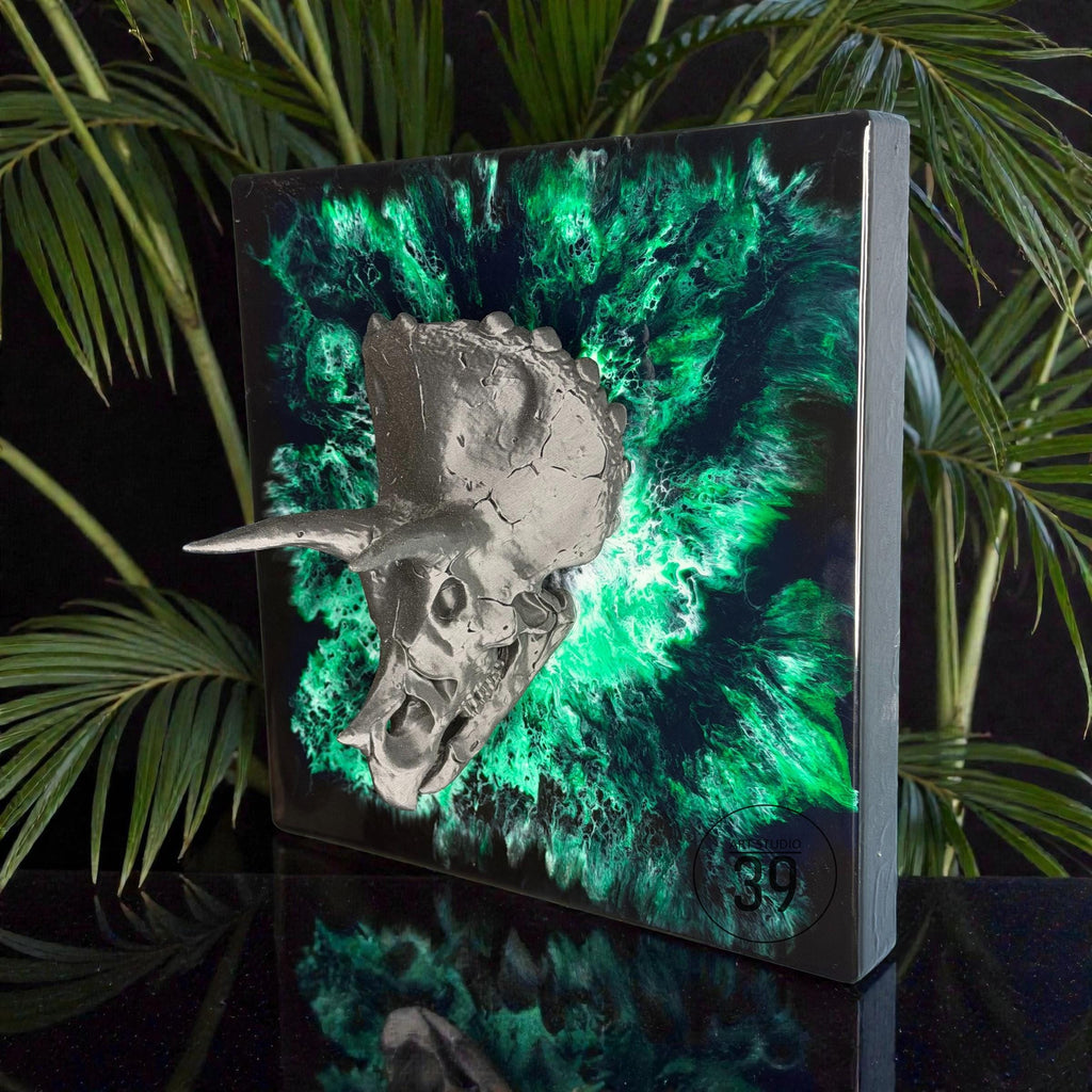 A metallic silver Triceratops skull sculpture mounted on a square black and green abstract background. The artwork has a textured appearance and is set against a backdrop of green foliage. The number "39" is visible on the lower right corner.