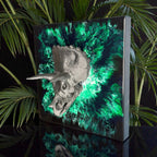 A metallic silver Triceratops skull sculpture mounted on a square black and green abstract background. The artwork has a textured appearance and is set against a backdrop of green foliage. The number "39" is visible on the lower right corner.