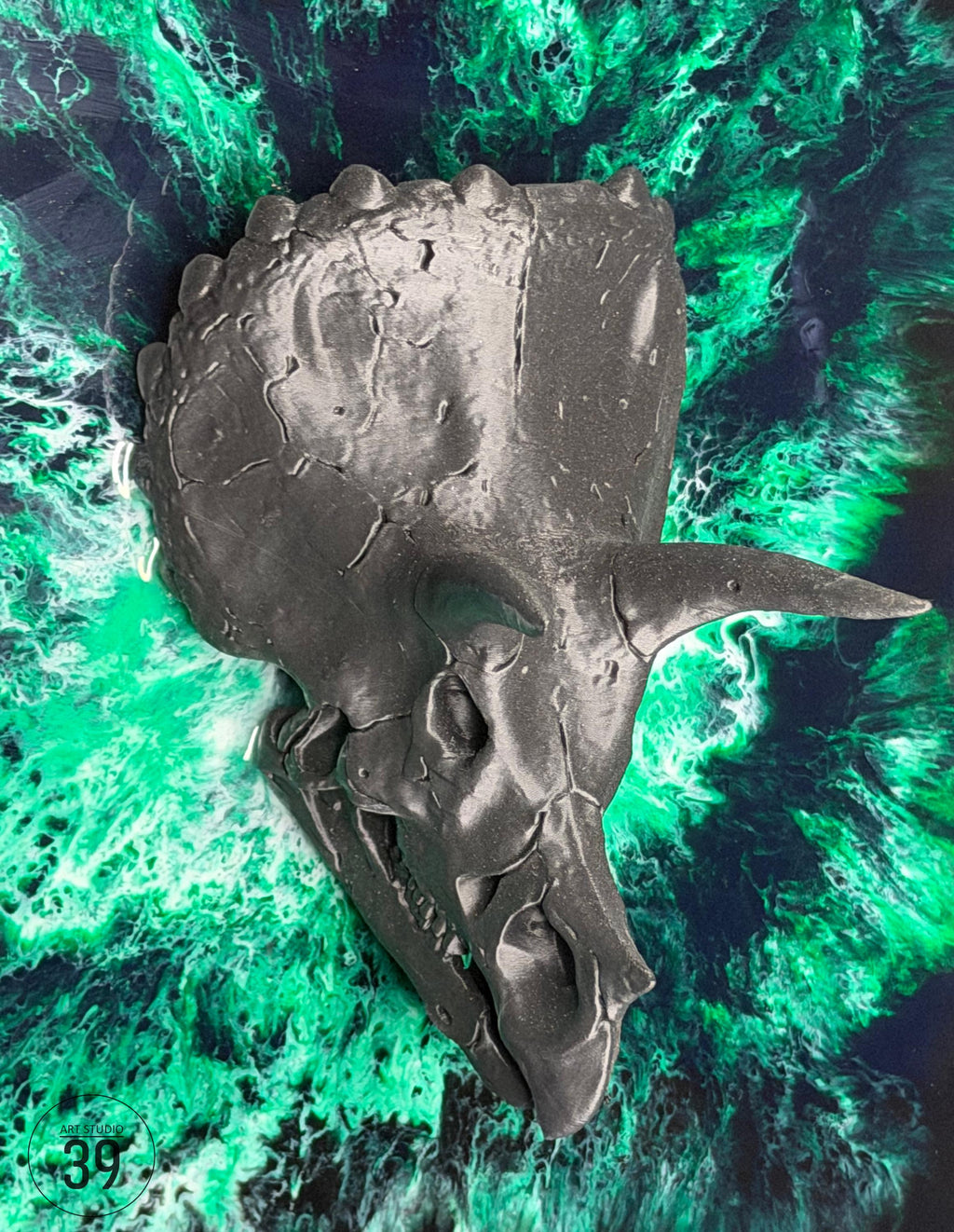 A dark gray triceratops skull sculpture with three horns and a detailed texture. The sculpture is set against a background of swirling green and black resin art.