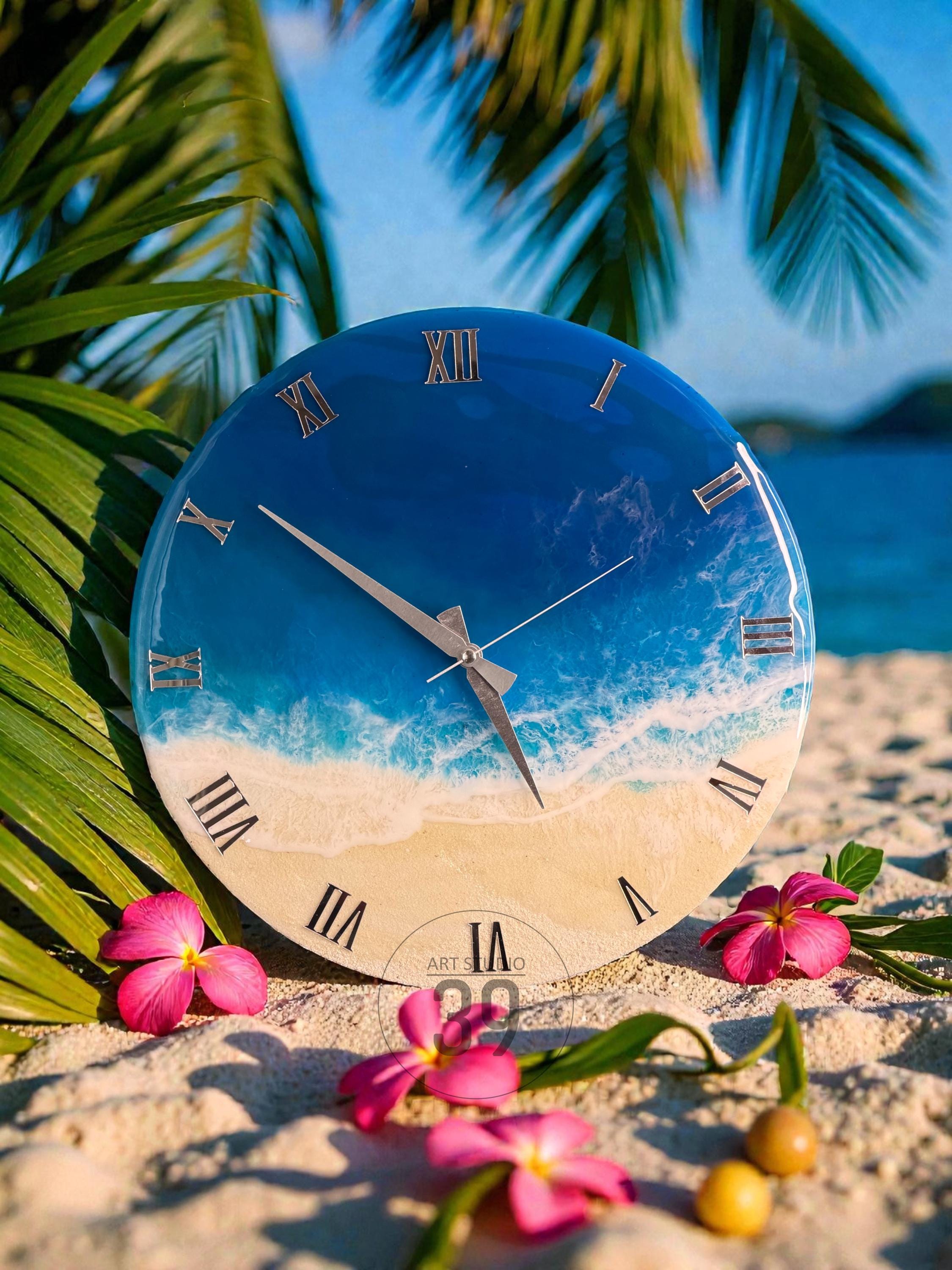 A round wall clock with a beach scene design. The clock face features a blue ocean, white waves, and a sandy beach. Roman numerals mark the hours. Pink flowers and palm leaves surround the clock.
