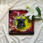 A square resin art piece with a dark gray dragon head sculpture. The background features a vibrant mix of red, yellow, and black colors. A white lily flower with green leaves is placed on the right side of the artwork.