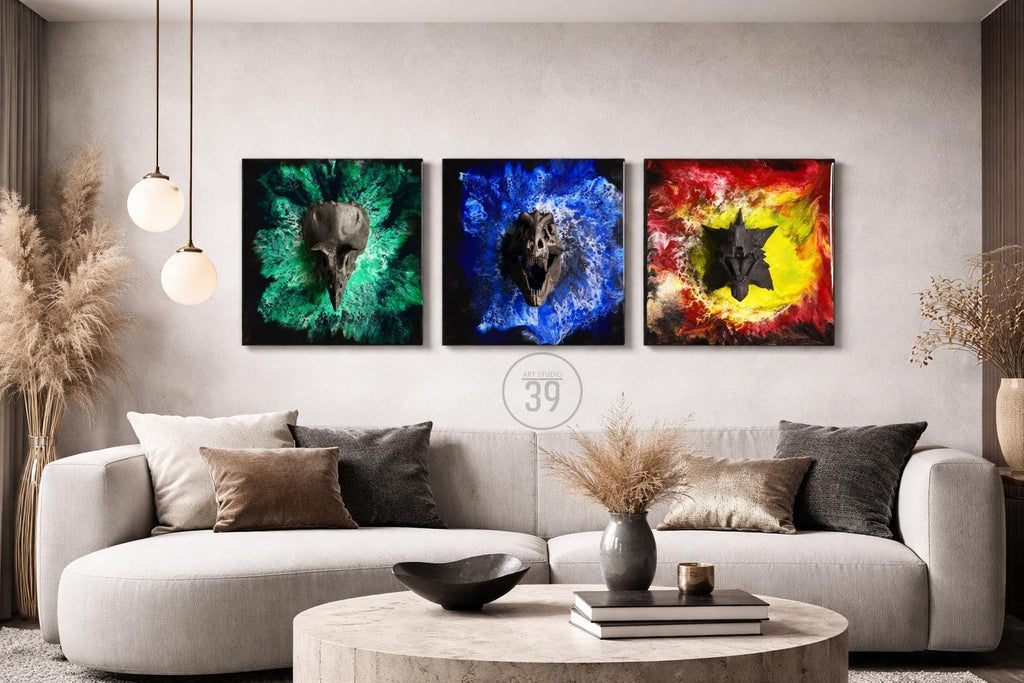 Three square wall art pieces featuring abstract skull designs. Each piece has a different color scheme: green, blue, and red/yellow. The artwork is displayed above a modern sofa in a living room setting.