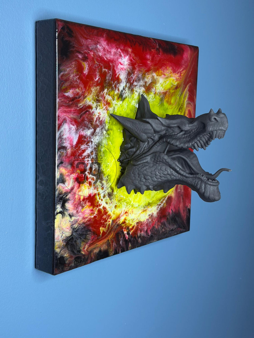 A square wall art piece featuring a 3D dragon head emerging from a vibrant, abstract background. The dragon head is gray, with detailed scales and an open mouth. The background has swirls of red, yellow, and black.