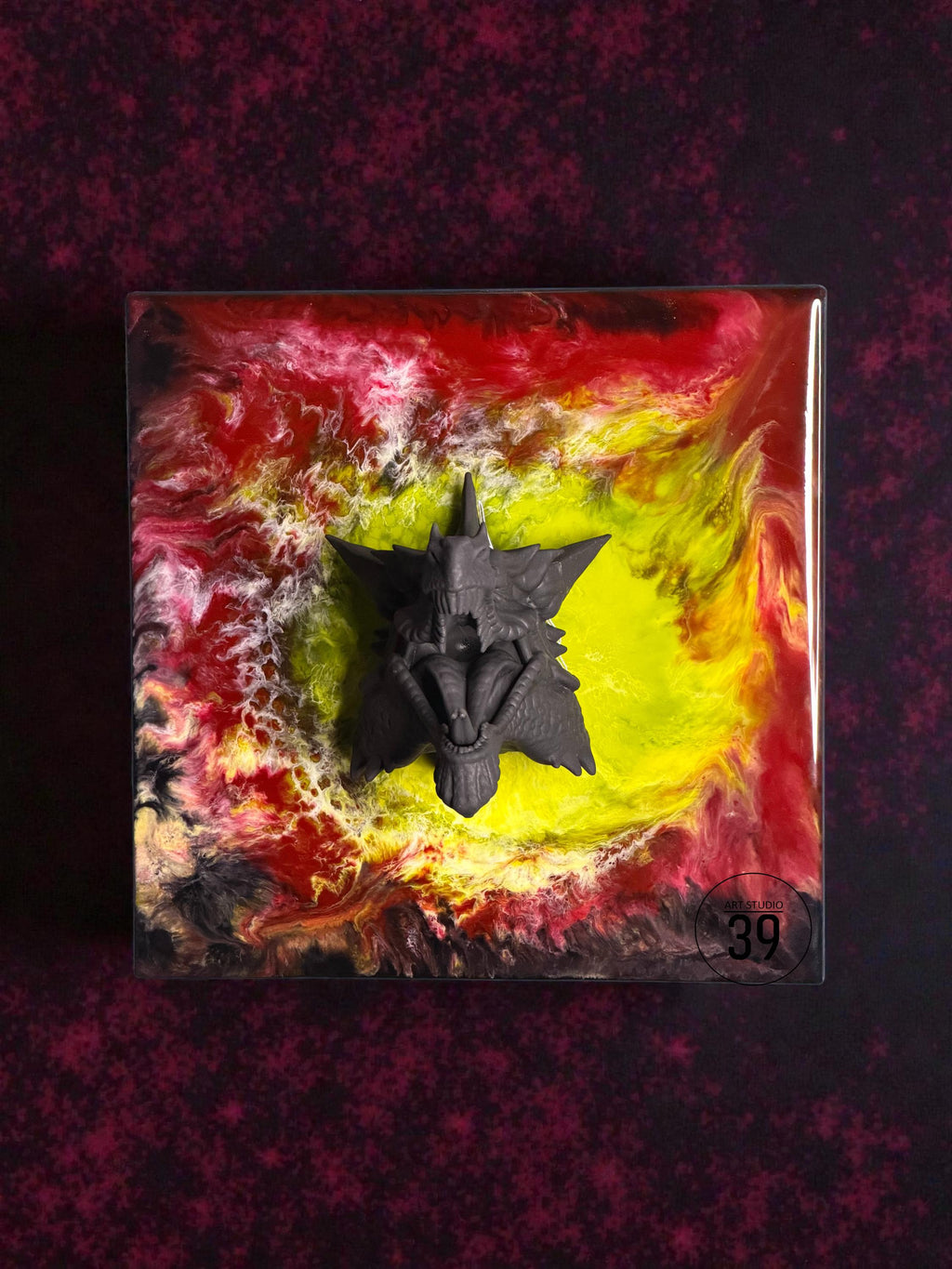 A square resin art piece featuring a dark gray, stylized creature sculpture. The background has a vibrant, swirling design in red, yellow, and black. The artwork is set against a dark, textured backdrop, with the text "ART STUDIO 39" visible.