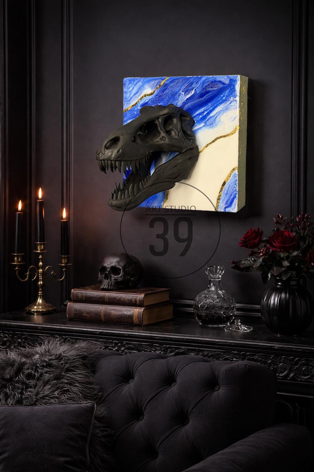 A black Tyrannosaurus Rex skull sculpture mounted on a canvas with blue and white abstract art and gold accents. The artwork is displayed above a black table with candles, books, and a skull.