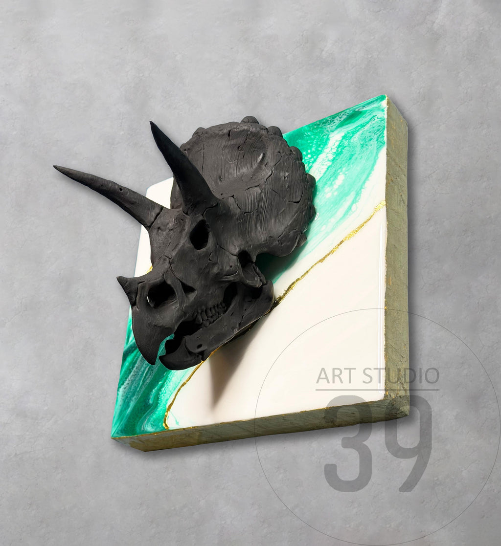 A black triceratops skull sculpture mounted on a square canvas. The canvas features a white and green marbled design with a gold accent. The artwork is labeled "ART STUDIO 39".