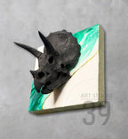 A black triceratops skull sculpture mounted on a square canvas. The canvas features a white and green marbled design with a gold accent. The artwork is labeled "ART STUDIO 39".