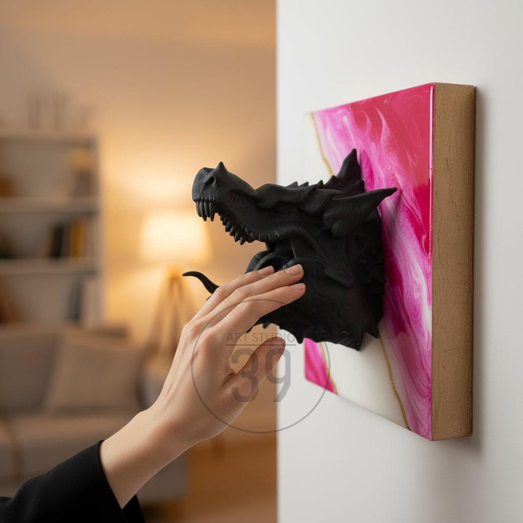 A black dragon head sculpture emerging from a pink and white abstract painting on a wooden frame. The dragon's head is detailed with sharp teeth and textured scales. The artwork is mounted on a white wall.