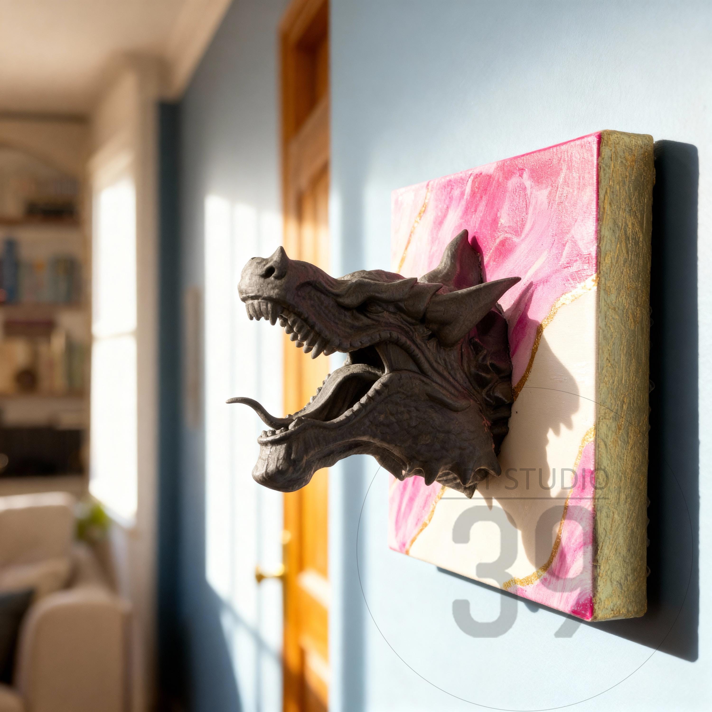 A dark gray dragon head sculpture with an open mouth, mounted on a pink and gold canvas. The sculpture is affixed to a wall, creating a 3D effect. The canvas has the text "STUDIO 39".