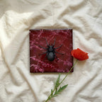 A square, dark red coaster with a black spider sculpture. A red flower and green stem are next to the coaster. The coaster has a marbled effect. The text "ART STUDIO 39" is in the lower left corner.