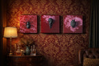 Three square wall art pieces with dark red backgrounds. Each features a black sculptural element: a spider, a bird skull, and a human heart. The art hangs on a patterned red and gold wallpaper.