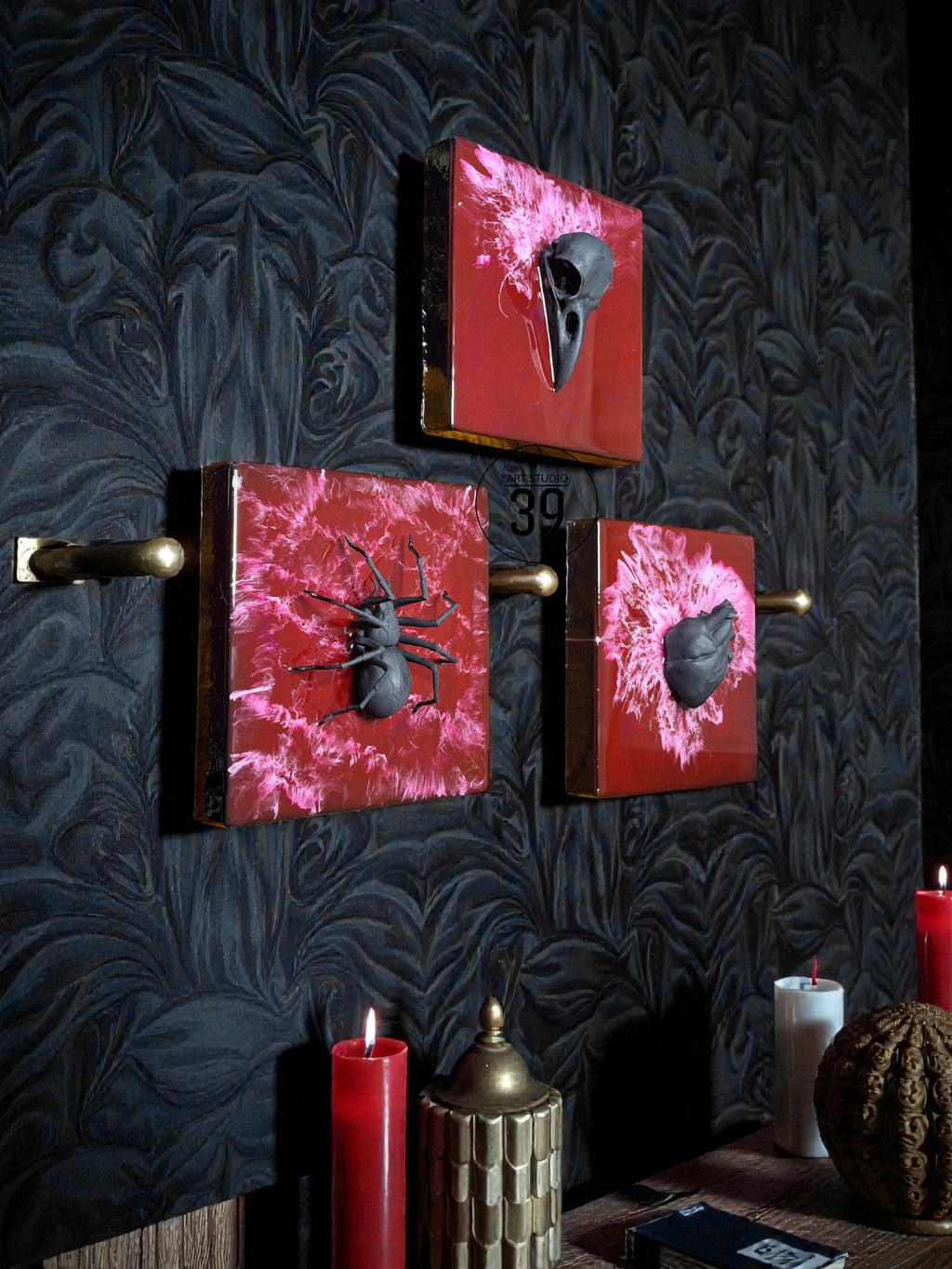 Three square wall art pieces with a red and pink marbled background, each featuring a dark gray object: a bird skull, a spider, and a heart-shaped object. The art is framed in gold and mounted on a dark patterned wall.