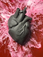 A dark gray, textured anatomical heart sculpture. The heart is detailed with visible veins and arteries. The background features a red and pink marbled design.