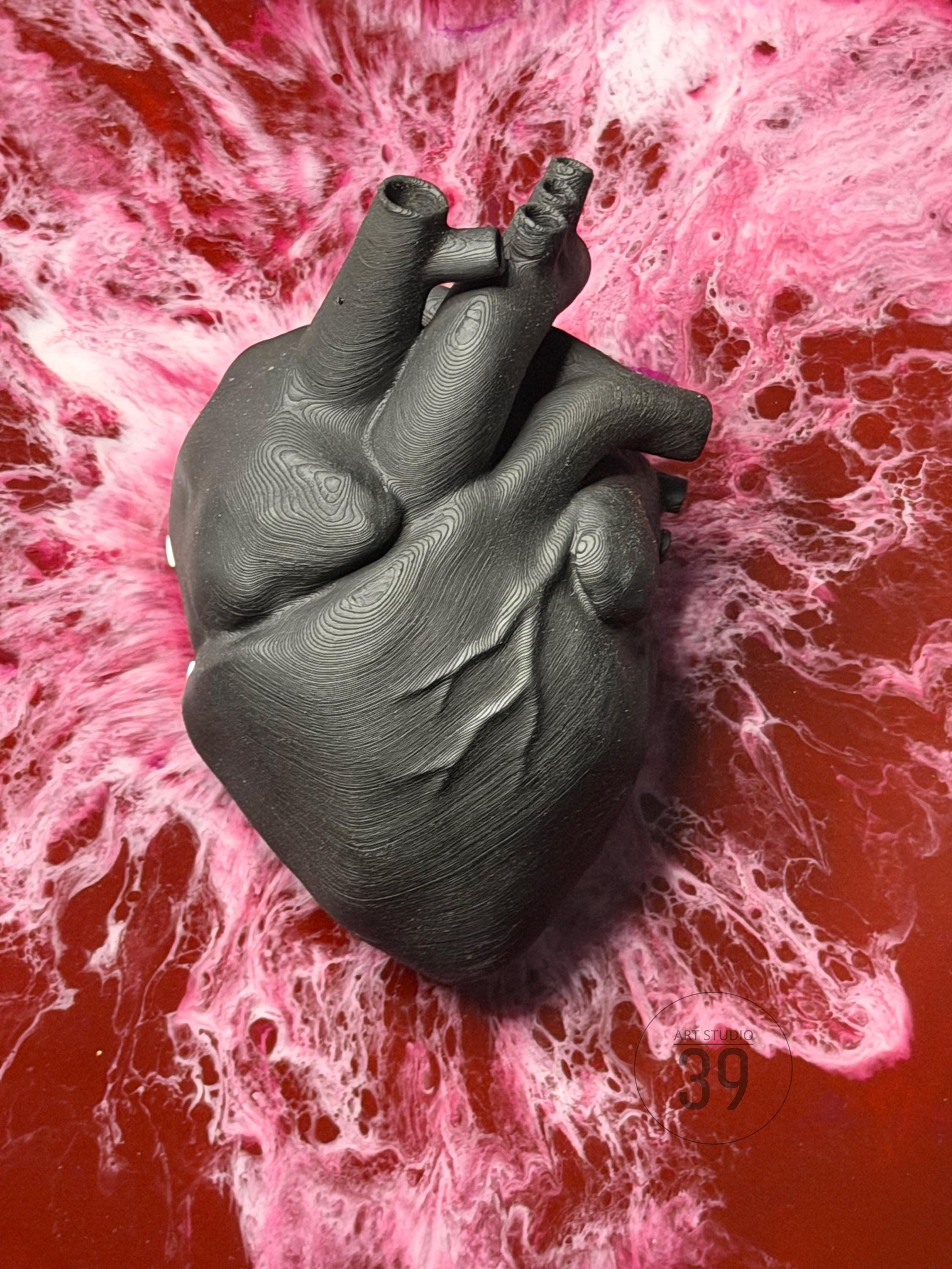 A dark gray, textured anatomical heart sculpture. The heart is detailed with visible veins and arteries. The background features a red and pink marbled design.