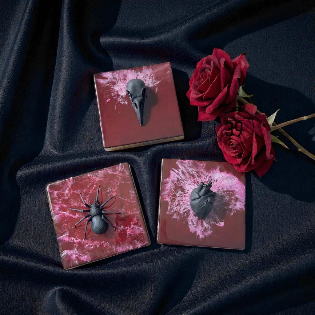 Three square coasters with dark red and pink abstract designs. Each coaster features a black sculptural element: a bird skull, a spider, and a human heart. Red roses are placed nearby on a dark blue fabric.