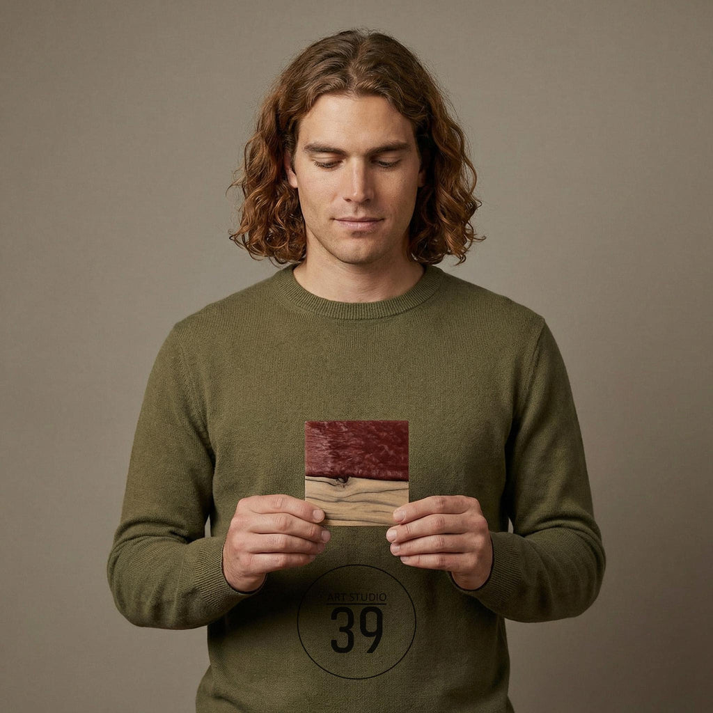 A square wooden coaster with a two-tone design. The top half is a deep red hue, while the bottom half is a natural wood color. The coaster is held in front of a person wearing a green sweater.