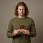 A square wooden coaster with a two-tone design. The top half is a deep red hue, while the bottom half is a natural wood color. The coaster is held in front of a person wearing a green sweater.
