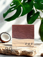 A square wooden block with a dark red resin top and a light wood base. The block is engraved with the initials "D &amp; Y" and the date "17.05.2023". A halved coconut and green leaves are in the background.