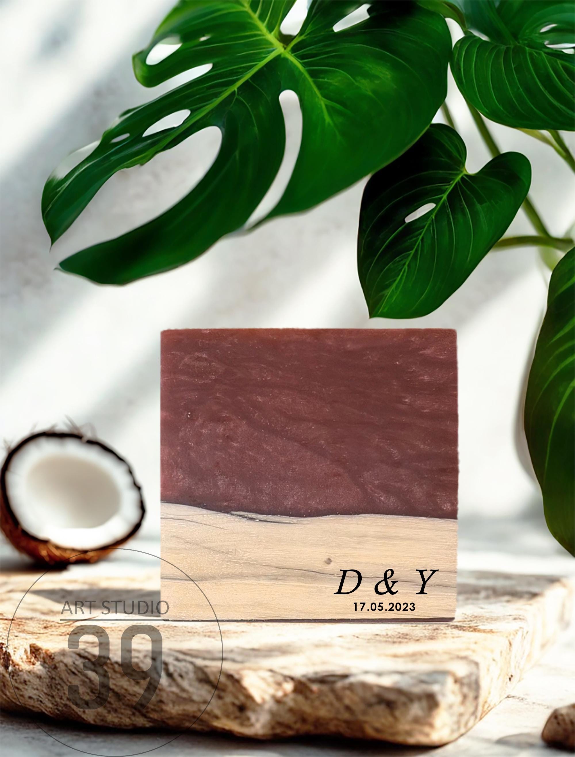 A square wooden block with a dark red resin top and a light wood base. The block is engraved with the initials "D &amp; Y" and the date "17.05.2023". A halved coconut and green leaves are in the background.
