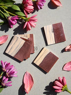Square wooden coasters with a two-tone design, featuring a light wood grain and a dark brown section. The coasters are arranged with pink flowers and leaves on a gray surface. The image includes the text "ST STUDIO 39".