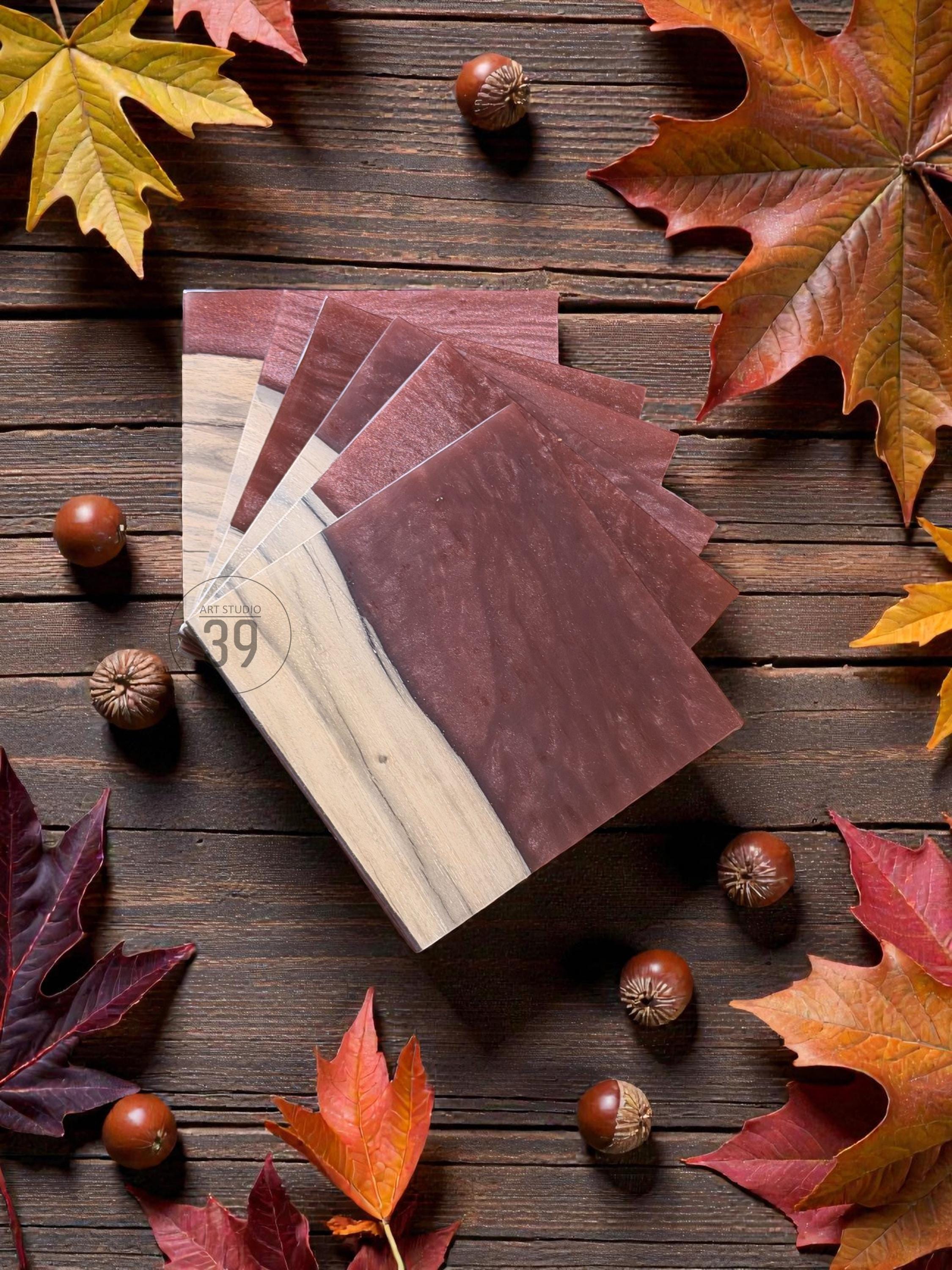 A stack of square wooden coasters with a natural wood grain and a deep burgundy resin inlay. The coasters are arranged on a dark wood surface, surrounded by colorful autumn leaves and acorns. The coasters have the text "ART STUDIO 39".