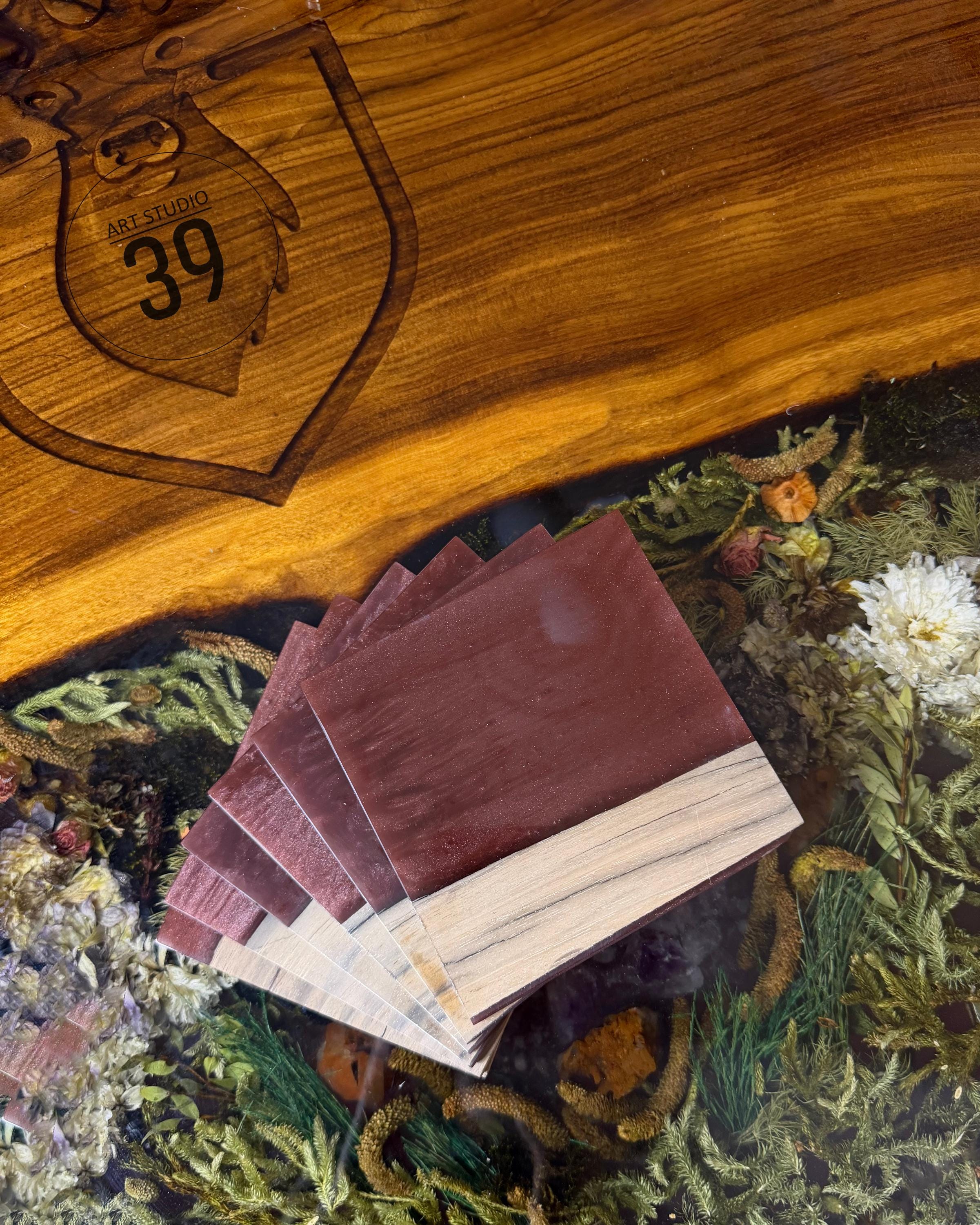 A stack of square, reddish-brown coasters with a light wood accent. The coasters are displayed on a wooden surface with the text "ART STUDIO 39" engraved. The background features dried flowers and greenery.