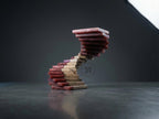 A wooden sculpture in a spiral staircase design. The steps alternate between natural wood and a reddish-brown resin. The sculpture has a modern aesthetic and is set against a dark gray background. The number "39" is in a circle on the sculpture.