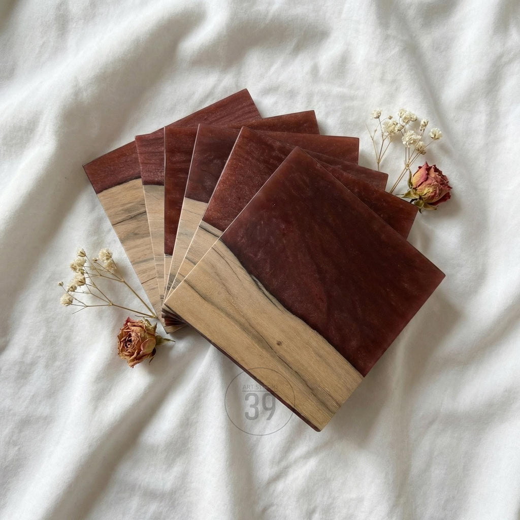 A stack of square coasters with a two-tone design. The top half is a deep burgundy color, while the bottom half is a light wood grain. The coasters are arranged on a white surface with dried flowers.