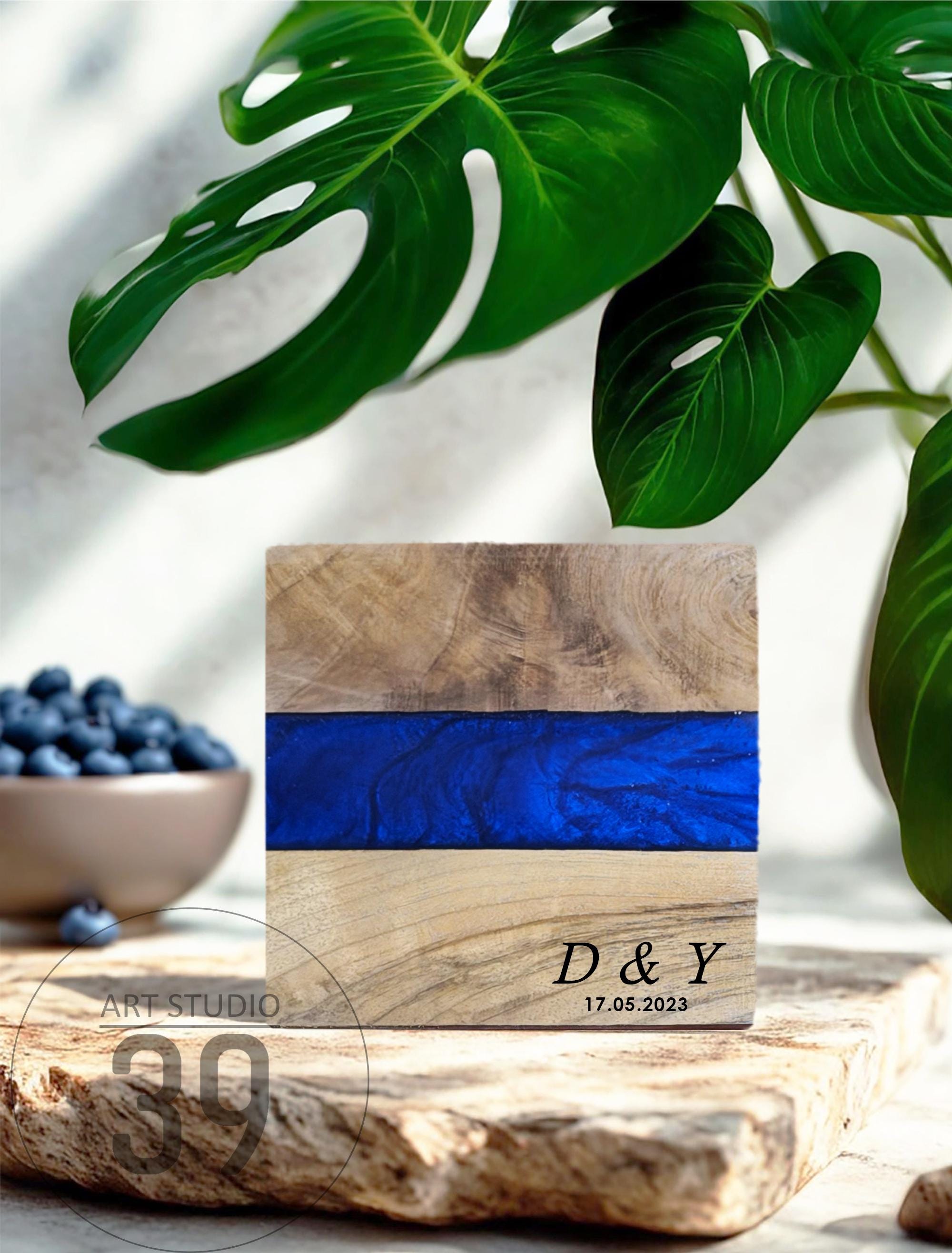 A wooden block with a vibrant blue resin stripe. The block is engraved with "D &amp; Y" and the date "17.05.2023". The background includes green leaves and a bowl of blueberries, suggesting a natural theme.
