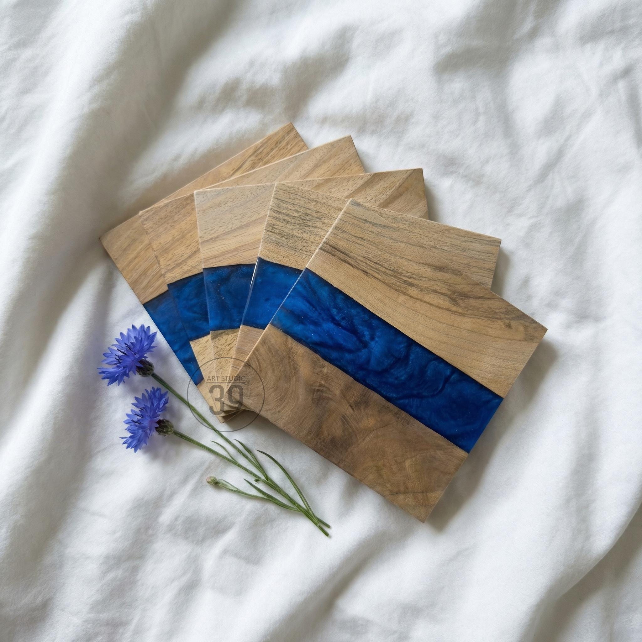 Set of wooden coasters with a blue resin stripe. The coasters are made of light-colored wood and have a rectangular shape. Two blue cornflower blossoms are placed near the coasters.