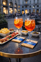 Two wine glasses filled with an orange cocktail and orange slice garnish. The glasses sit on wooden coasters with blue resin accents. Snacks and appetizers are arranged on a table.