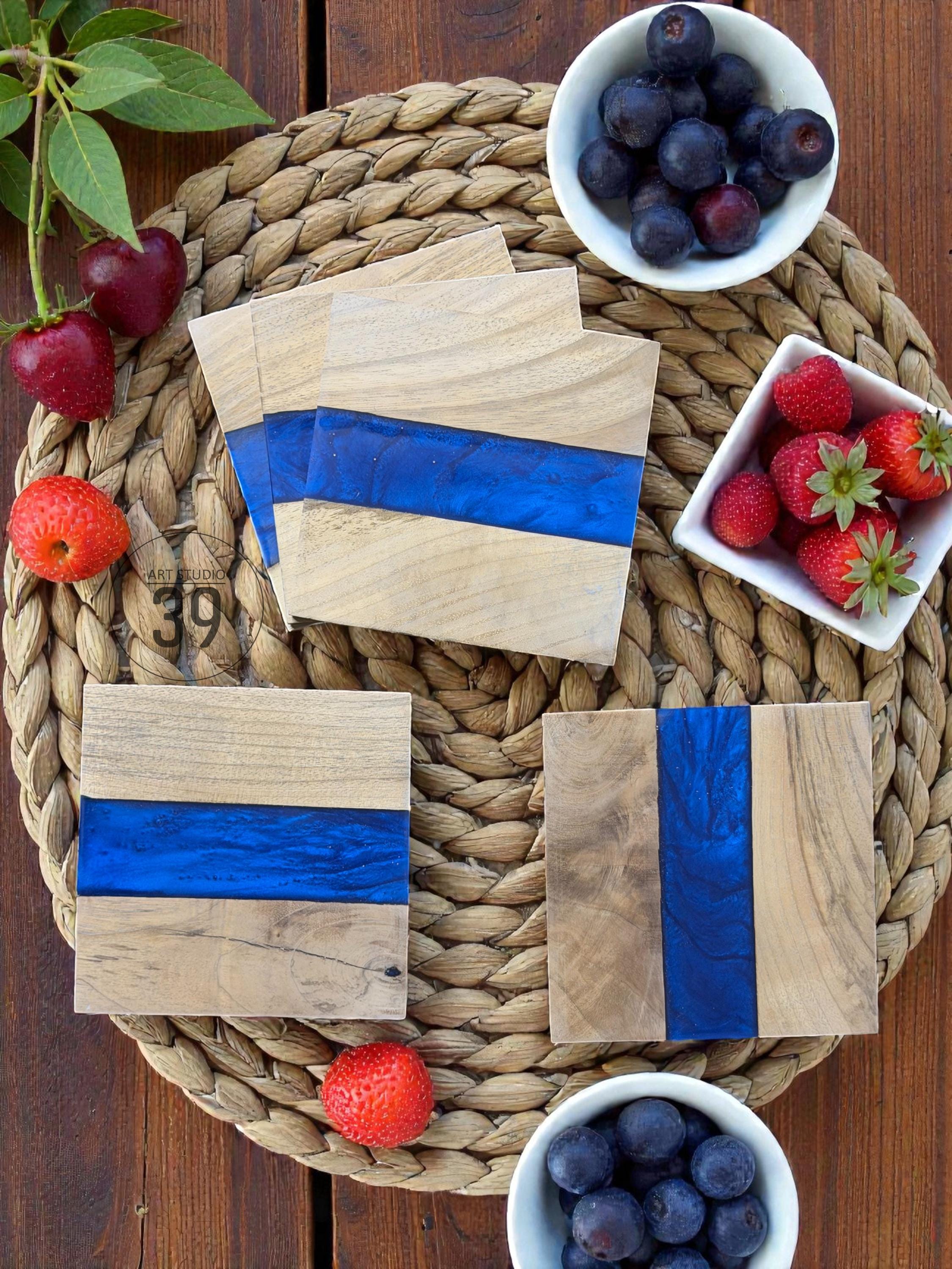 Wooden square coasters with a vibrant blue resin stripe. The coasters are arranged on a woven mat, accompanied by fresh strawberries, blueberries, and cherries. The wood grain is visible, adding a natural touch to the design.