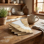 A stack of square wooden coasters with a white resin inlay, arranged on a rustic wooden table. A ceramic mug with a hot beverage sits nearby, with a small potted plant and kitchenware in the background. The coasters have the text "ART STUDIO 39".