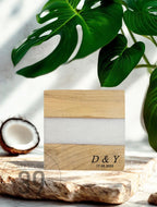 A square wooden block with a white resin stripe. The block has the initials "D &amp; Y" and the date "17.05.2023" engraved on it. The block is on a wooden surface, with a coconut half and green leaves in the background.