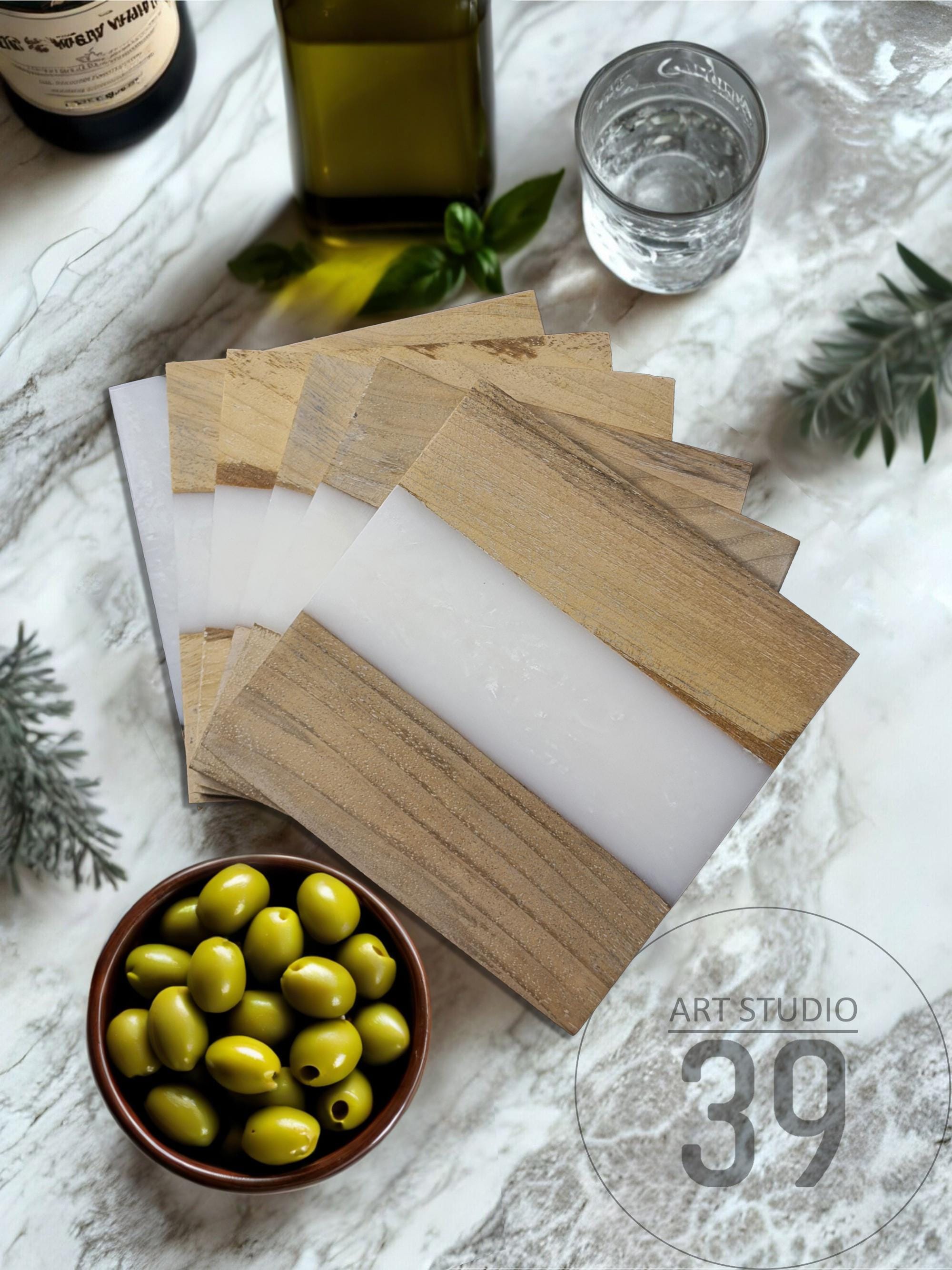 A collection of wooden and white resin coasters arranged on a marble surface. A small bowl of green olives, a glass of water, and bottles of oil and wine are also present. The coasters have a rectangular shape and are made of wood and white resin.