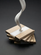 A stack of wooden coasters with a white resin inlay. A white, flowing sculpture appears to pour onto the top coaster. The coasters have a natural wood grain finish. The image has a modern, artistic aesthetic.