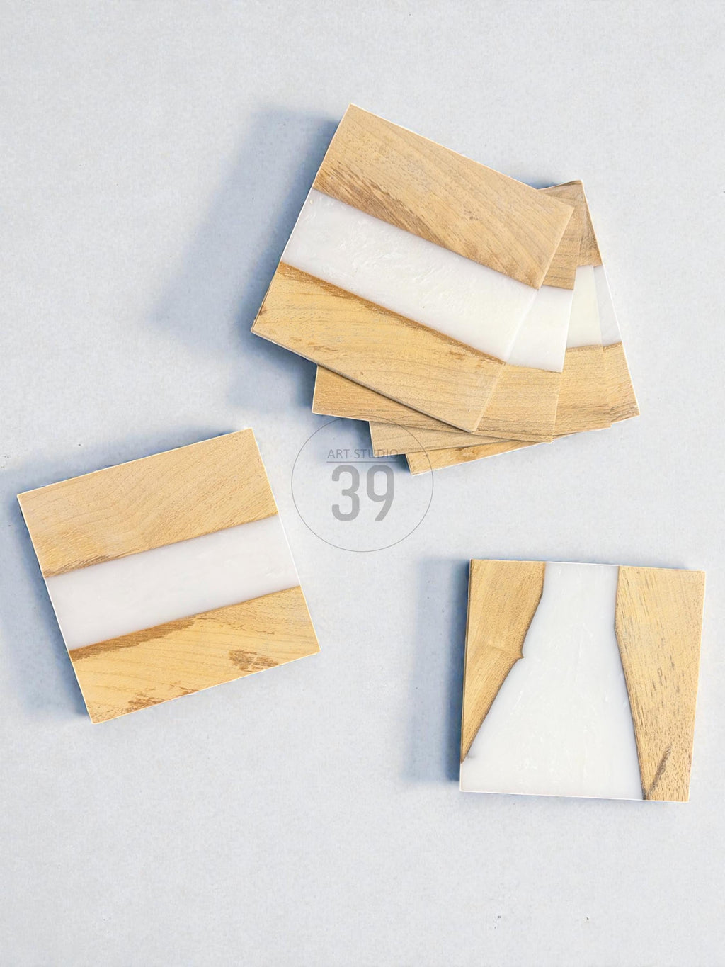 Wooden square coasters with a white resin stripe. The coasters are made of light-colored wood and have a smooth, glossy finish. The coasters are arranged on a light blue surface.